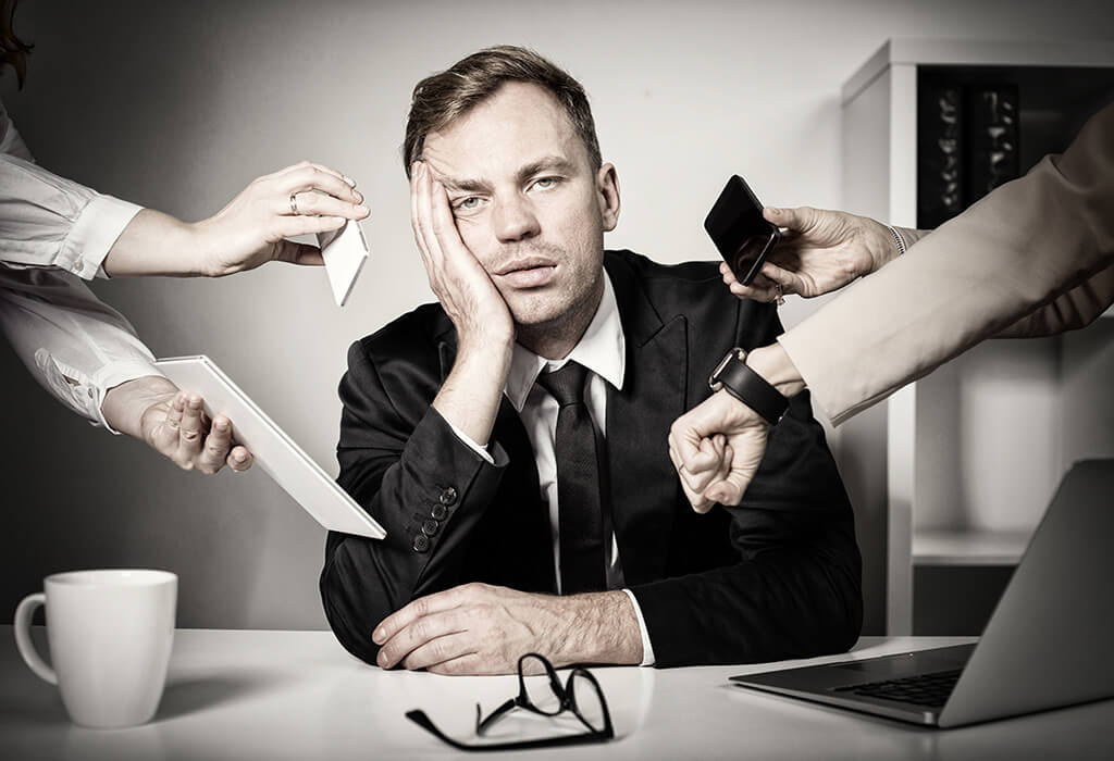 Overwhelmed manager at a desk as hands thrust phones and documents at him from every side